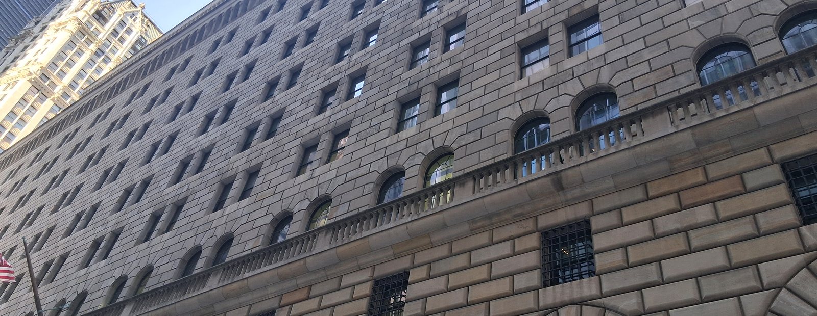 The limestone facade of the Federal Reserve Bank of New York, seen from the street, with its arched windows and iron lanterns.