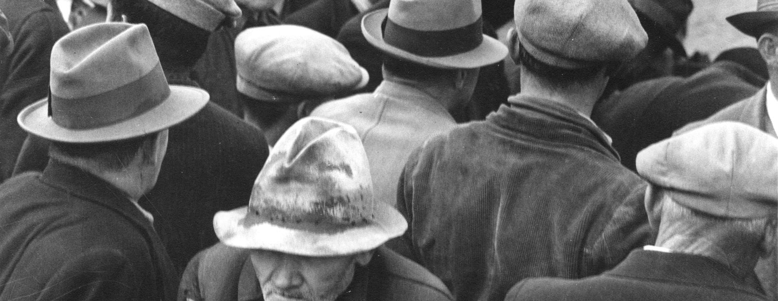 A breadline in San Francisco in 1932. Men in hats face away from the camera; one turns toward it, unshaven, holding a tin cup.